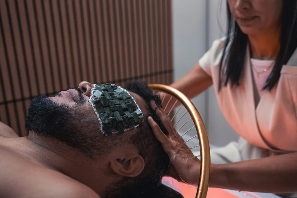 Terapeuta durante um ritual de Head Spa, com o cliente deitado, máscara de pedras sobre os olhos e o arco de cobre